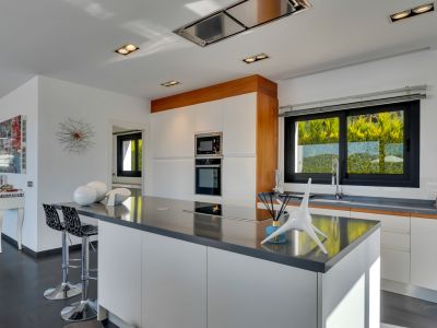 Cuisine moderne avec grand îlot central à plaque intégrée, meubles blancs sans poignées et électroménager encastré. Grande fenêtre offrant lumière naturelle et vue sur la verdure.