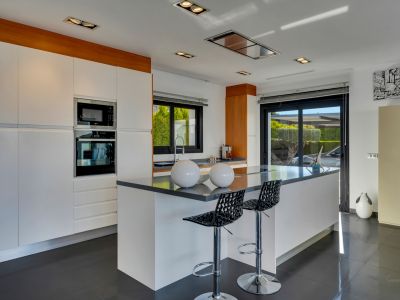 Cuisine moderne avec grand îlot central et sièges, électroménager intégré, armoires blanches et accès direct à l'extérieur par porte coulissante vitrée.