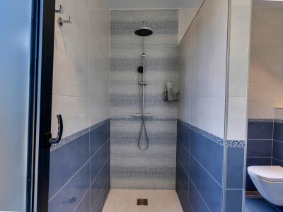Douche à l'italienne carrelée de bleu et blanc avec une bande décorative en mosaïque. Comprend une pomme de douche pluie et une douchette, séparée par des parois vitrées.