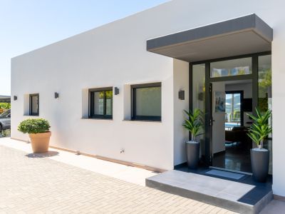 Entrée moderne d'une villa blanche avec porte vitrée encadrée de noir et auvent gris. Extérieur pavé, fenêtres à volets et parking couvert visible.
