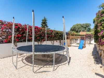 Espace de jeux extérieur avec trampoline et structure en bois avec toboggan bleu sur gravier. Des bougainvilliers luxuriants et un mur de pierre avec des vignes violettes encadrent la zone sous un ciel clair.