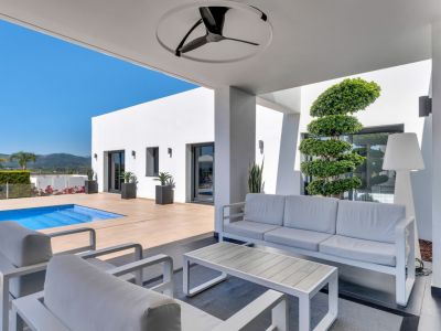 Espace lounge extérieur couvert avec mobilier blanc contemporain, ventilateur de plafond et accès direct à la piscine. Architecture minimaliste et vue sur les montagnes.