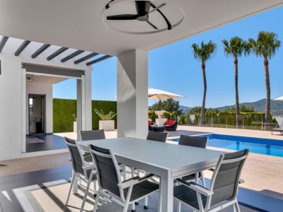 Espace repas extérieur couvert avec table et chaises modernes, ventilateur de plafond, vue sur la piscine, les palmiers et les montagnes. Une salle de bain extérieure est visible.