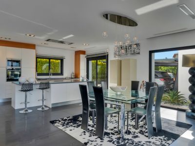 Espace repas intégré à une cuisine moderne blanche, avec table en verre, chaises noires et lustre contemporain. De grandes portes coulissantes mènent à l'extérieur.