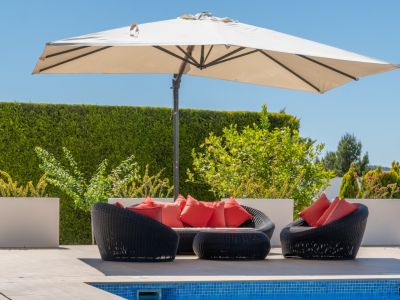 Espace salon extérieur avec mobilier en rotin noir et coussins rouges vifs, ombragé par un grand parasol, à côté de la piscine carrelée.