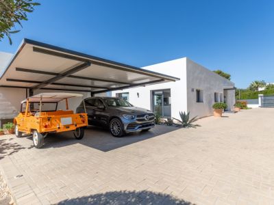 Extérieur de villa moderne avec double abri de voiture couvert à structure sombre, abritant un SUV Mercedes et une voiture orange décapotable, sur une allée pavée.