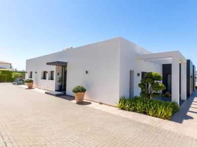 Extérieur de villa moderne avec façade blanche épurée, allée pavée et aménagement paysager minimaliste. Arbre sculpté, pergola et collines lointaines.