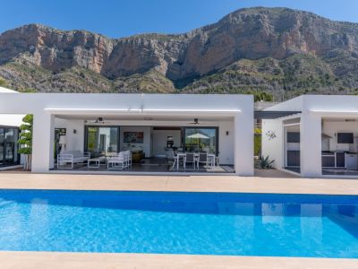 La villa moderne offre une piscine bleue lumineuse, une terrasse couverte avec salon et salle à manger extérieurs, et une cuisine d'été intégrée, face au Montgó.