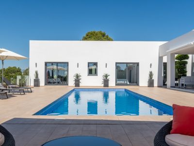Piscine rectangulaire en mosaïque bleue sur une terrasse carrelée, devant une façade blanche moderne avec de grandes portes coulissantes et un salon extérieur couvert.