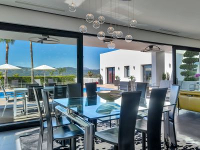 Salle à manger moderne avec table en verre et chaises noires, s'ouvrant sur une terrasse piscine avec transats, palmiers et vue sur les montagnes.