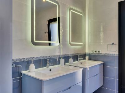 Salle de bain contemporaine avec double vasque suspendue bleu clair, plans de travail blancs et deux miroirs LED au-dessus de murs carrelés bleus et gris.