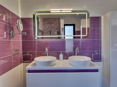 Salle de bain moderne avec double vasque, carrelage mural violet horizontal et miroir lumineux reflétant la douche en mosaïque.