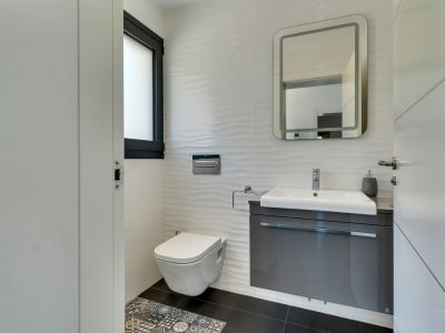 Salle de bain moderne avec toilettes suspendues, meuble vasque gris flottant et carrelage mural blanc texturé. Un miroir éclairé et des carreaux de sol décoratifs complètent l'espace.