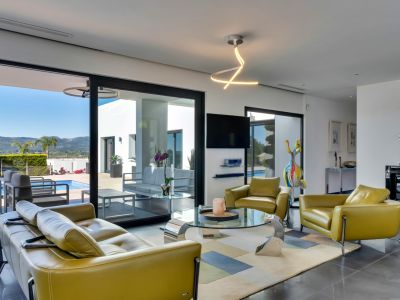Salon lumineux avec canapé et fauteuils en cuir vert olive, table basse en verre et accès direct à la terrasse, piscine et vue sur la montagne.