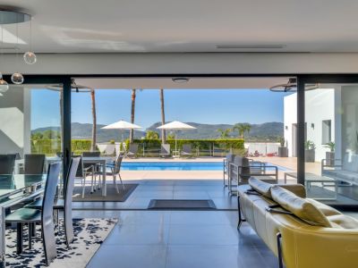 Salon-salle à manger ouvert avec table en verre, suspensions modernes et canapé jaune, s'ouvrant sur la terrasse de la piscine avec transats et vues lointaines sur les montagnes.