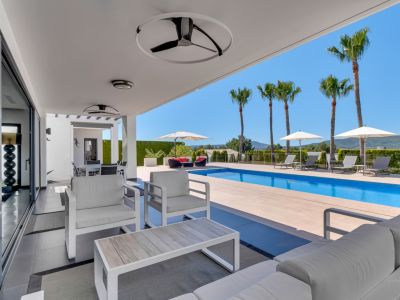 Terrasse couverte moderne avec mobilier de salon blanc, ventilateurs de plafond et grandes baies vitrées, s'ouvrant sur une piscine rectangulaire et des vues lointaines.