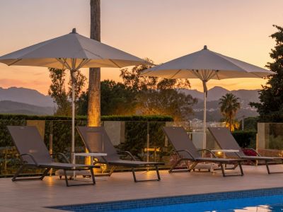 Terrasse de piscine au crépuscule avec quatre chaises longues grises, deux parasols éclairés et une balustrade en verre, offrant des vues sur les montagnes.