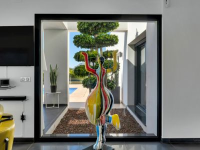 Une sculpture moderne et colorée encadre la vue depuis le salon à travers une grande fenêtre noire, donnant sur un jardin paysager avec un arbre unique et une piscine.