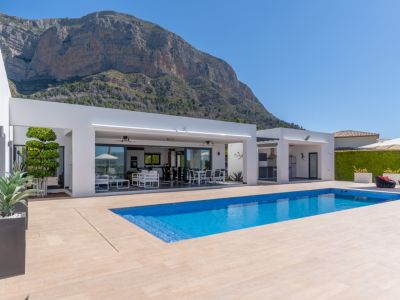 Vaste espace extérieur avec piscine rectangulaire en mosaïque bleue, terrasse carrelée claire et villa blanche moderne avec salon et salle à manger couverts intégrés, face au Montgó.