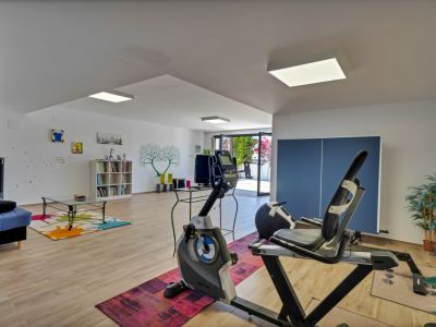 Vaste pièce polyvalente au niveau inférieur, équipée d'appareils de fitness, d'un coin salon avec canapé bleu et d'une bibliothèque. Accès direct à l'extérieur et parquet clair.