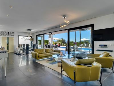 Vaste salon-salle à manger décloisonné avec accès direct à la terrasse et piscine chauffée via de grandes baies vitrées. Mobilier vert olive et sol carrelé gris foncé.