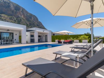 Vaste terrasse de piscine carrelée de bleu, espace barbecue extérieur et chaises longues sous parasols, avec vue sur l'imposant Montgó.