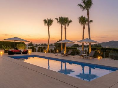 Vaste terrasse de piscine extérieure avec chaises longues élégantes, palmiers illuminés et coin salon, encadrée par un coucher de soleil vibrant et des vues naturelles.