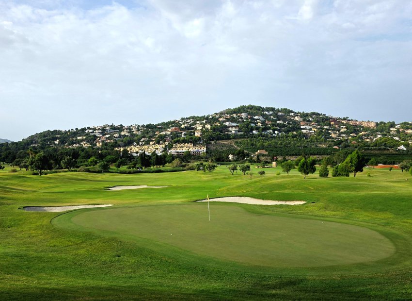 Vaste green de golf avec bunkers de sable et drapeau, adossé à une colline verdoyante parsemée de résidences de luxe sous un ciel nuageux.