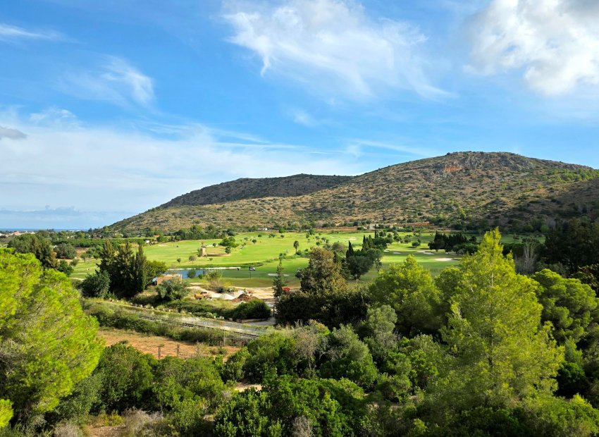 Vue panoramique sur un parcours de golf verdoyant avec plan d'eau et tour de pierre distinctive, encadré par une montagne aride et la mer lointaine.