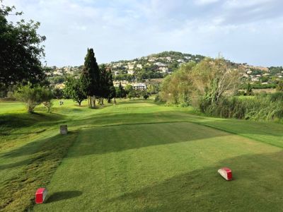 Un départ de golf méticuleusement entretenu s'ouvre sur un fairway verdoyant, bordé de cyprès et de résidences à flanc de colline. Des repères rouges indiquent le début du parcours.