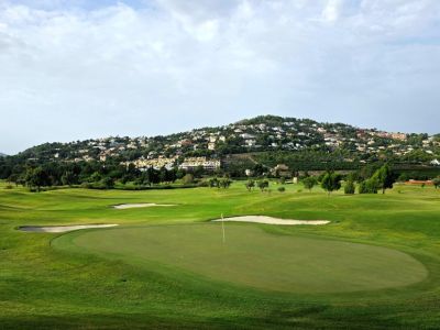Vaste green de golf avec bunkers de sable et drapeau, adossé à une colline verdoyante parsemée de résidences de luxe sous un ciel nuageux.