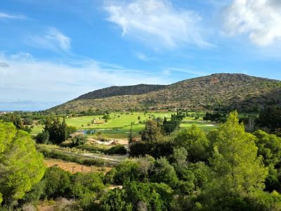 Vue panoramique sur un parcours de golf verdoyant avec plan d'eau et tour de pierre distinctive, encadré par une montagne aride et la mer lointaine.