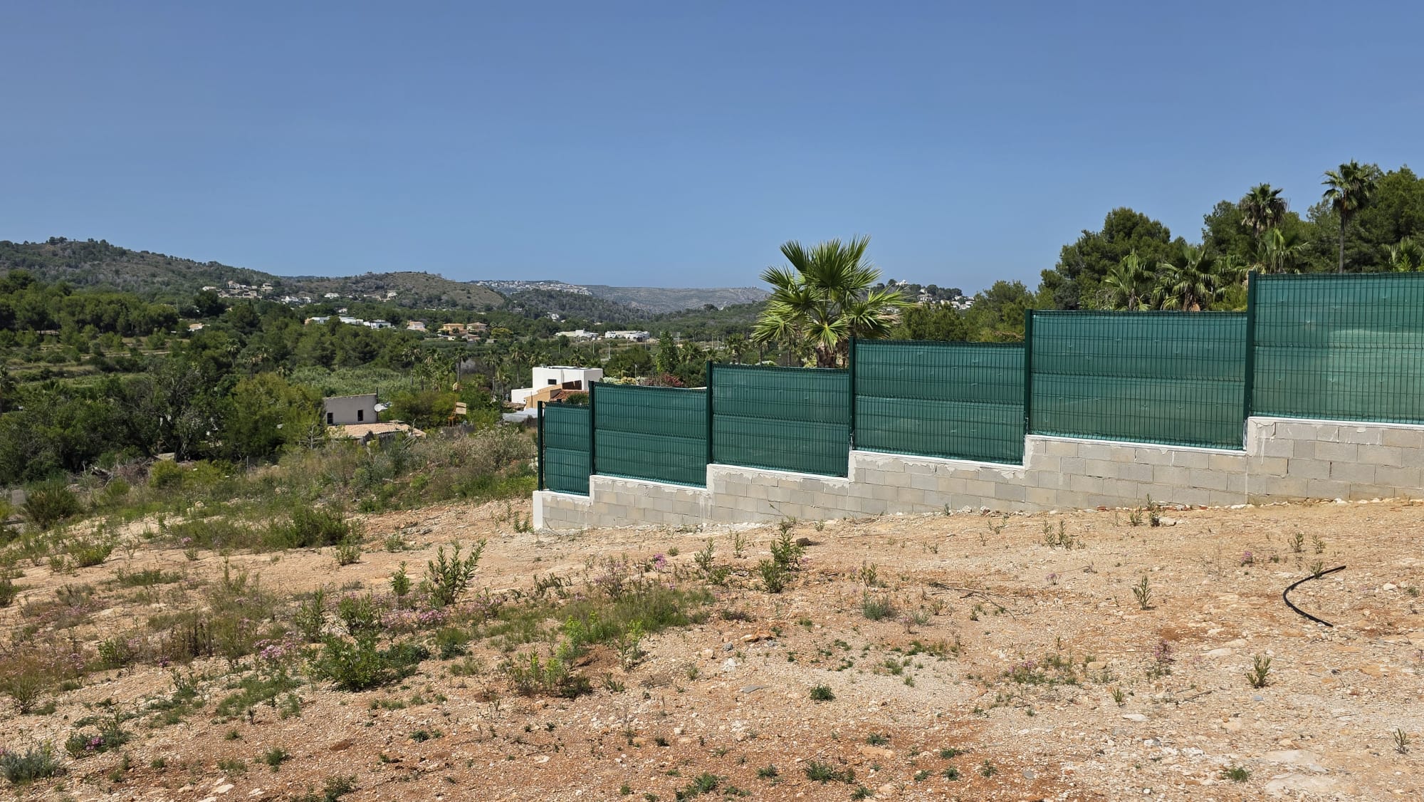 Terrain aride avec clôture étagée en blocs de béton et panneaux grillagés verts. Vues étendues sur des collines méditerranéennes parsemées de résidences sous un ciel clair. Terrain aride avec clôture étagée en blocs de béton et panneaux grillagés verts. Vues étendues sur des collines méditerranéennes parsemées de résidences sous un ciel clair.