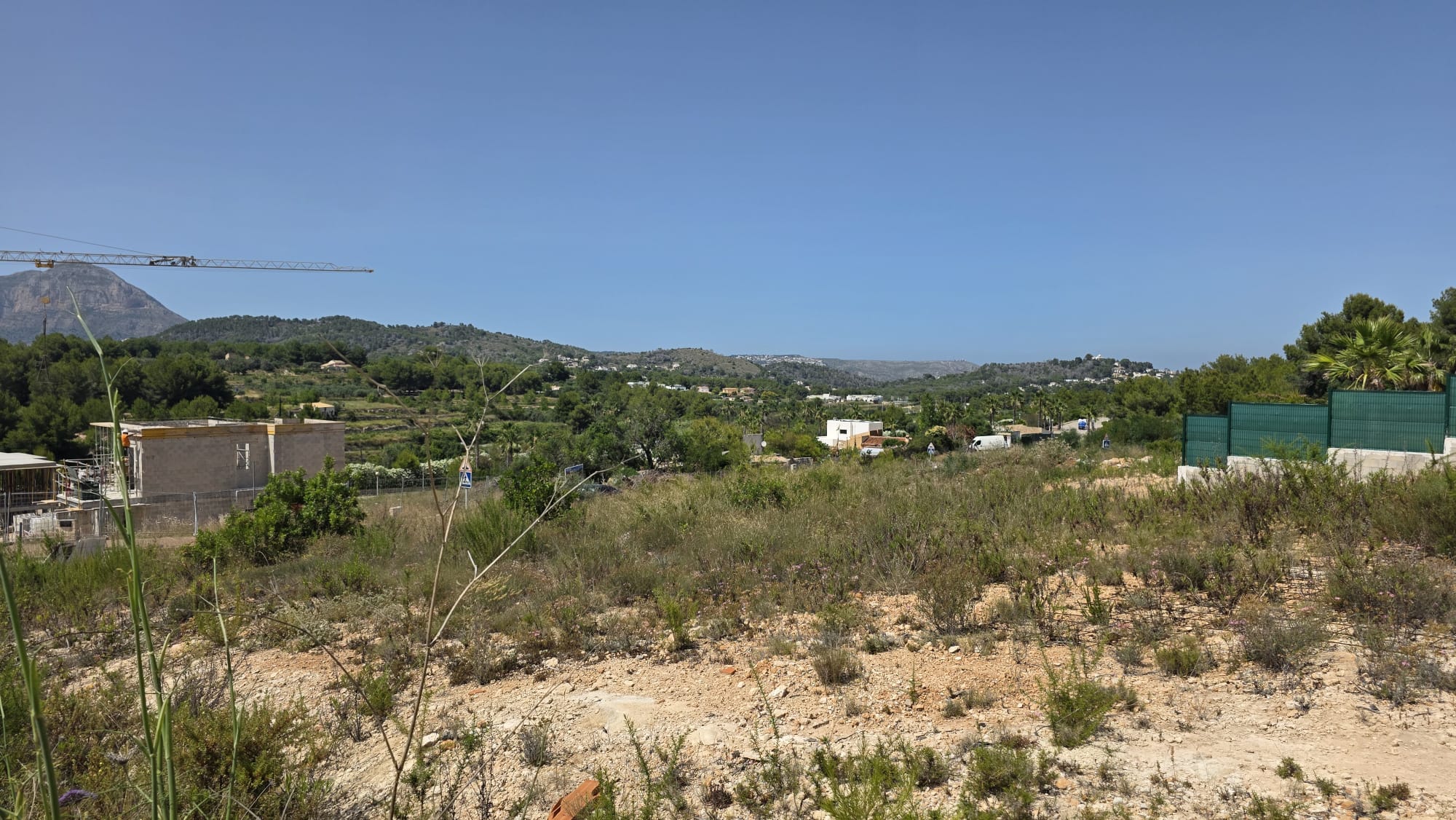 Terrain avec végétation naturelle et sol aride, encadré par des collines verdoyantes lointaines et des zones résidentielles. Une grue de construction et un bâtiment en cours sont visibles. Terrain avec végétation naturelle et sol aride, encadré par des collines verdoyantes lointaines et des zones résidentielles. Une grue de construction et un bâtiment en cours sont visibles.