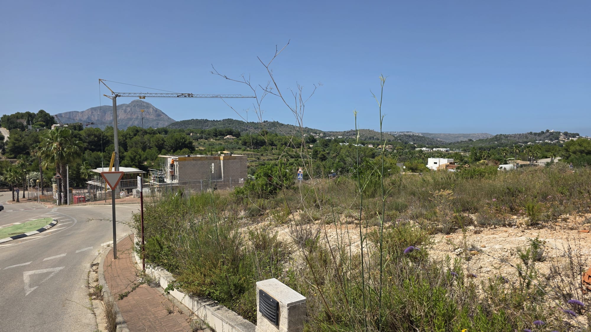 Terrain non aménagé avec végétation naturelle, un carrefour routier pavé et un chantier de construction lointain avec grue. Des vues sur la montagne majestueuse sous un ciel bleu clair. Terrain non aménagé avec végétation naturelle, un carrefour routier pavé et un chantier de construction lointain avec grue. Des vues sur la montagne majestueuse sous un ciel bleu clair.