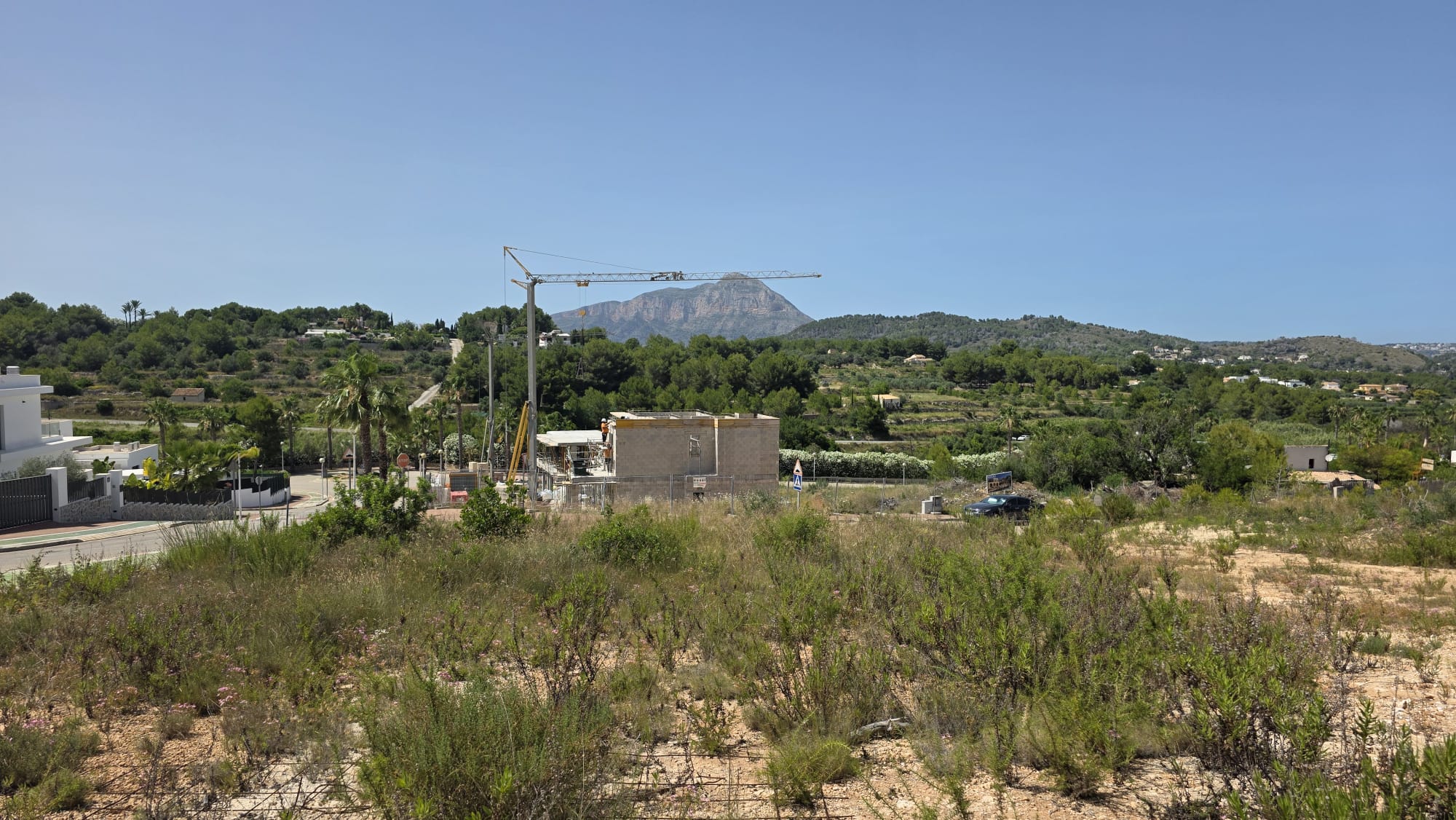Vaste terrain avec végétation naturelle et ferraillage visible, surplombant un chantier avec grue, des collines verdoyantes et le majestueux Montgó sous un ciel bleu clair. Vaste terrain avec végétation naturelle et ferraillage visible, surplombant un chantier avec grue, des collines verdoyantes et le majestueux Montgó sous un ciel bleu clair.