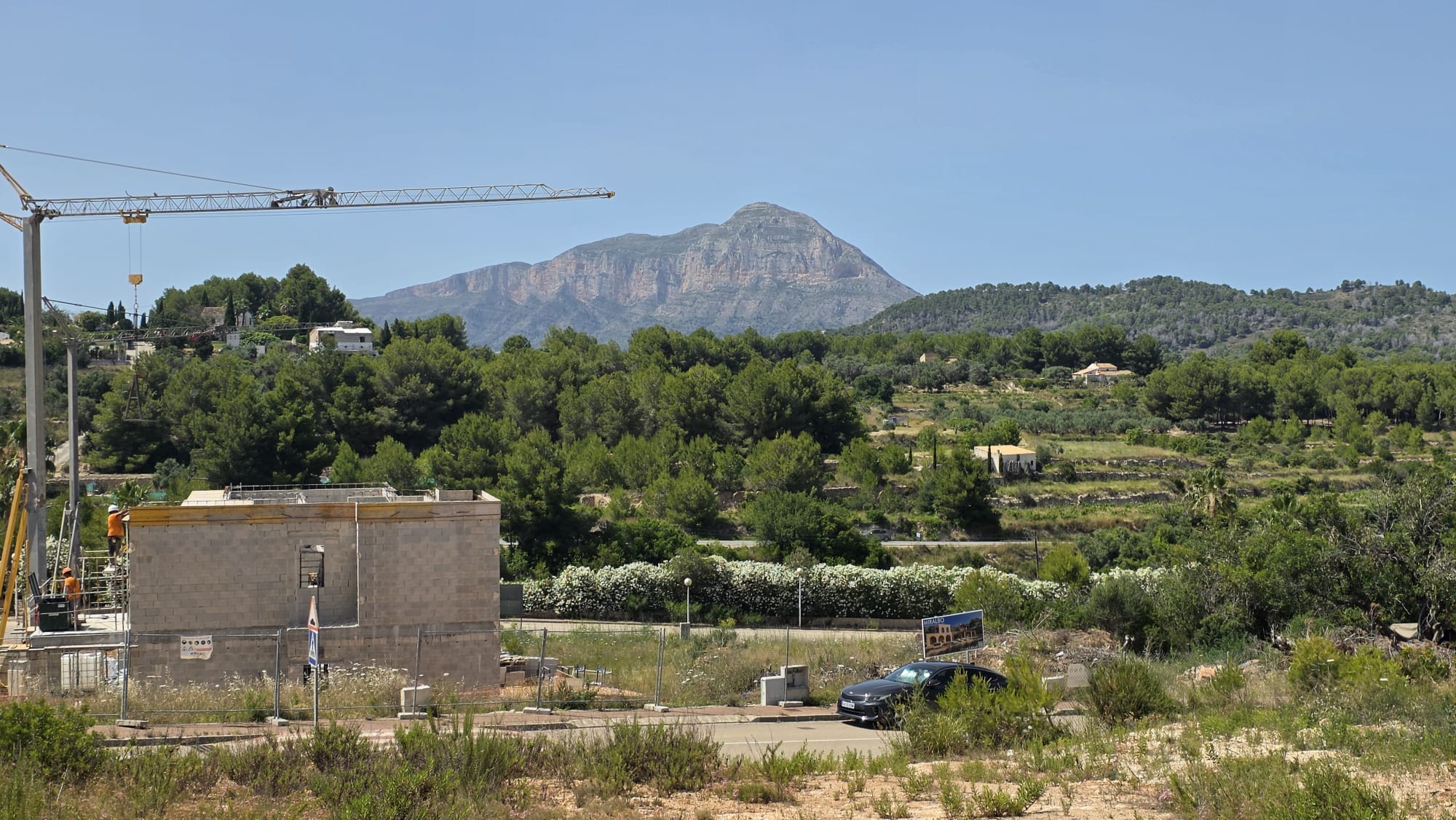Vue d'un chantier avec une structure en blocs de béton et une grue, encadré par une végétation méditerranéenne luxuriante, des collines en terrasses et la montagne du Montgó sous un ciel clair. Vue d'un chantier avec une structure en blocs de béton et une grue, encadré par une végétation méditerranéenne luxuriante, des collines en terrasses et la montagne du Montgó sous un ciel clair.