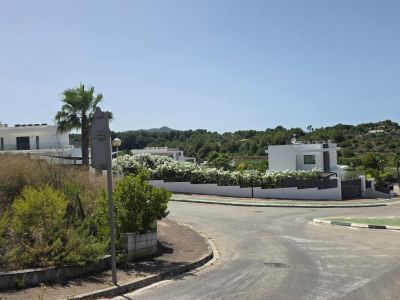 Scène de rue résidentielle montrant des villas blanches modernes avec balcons vitrés, entourées de flore méditerranéenne et de collines verdoyantes. Infrastructures routières et ciel clair.