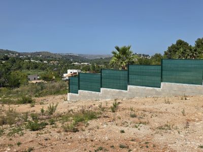 Terrain aride avec clôture étagée en blocs de béton et panneaux grillagés verts. Vues étendues sur des collines méditerranéennes parsemées de résidences sous un ciel clair.