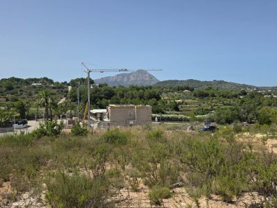 Vaste terrain avec végétation naturelle et ferraillage visible, surplombant un chantier avec grue, des collines verdoyantes et le majestueux Montgó sous un ciel bleu clair.