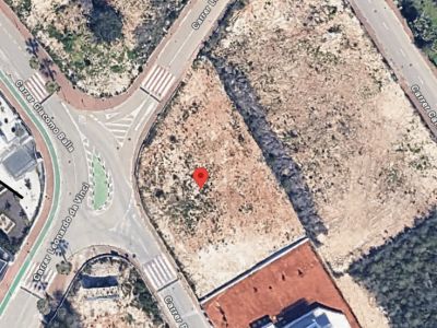 Vue aérienne d'une parcelle urbaine non bâtie, marquée d'une épingle rouge, située à côté d'un carrefour avec un rond-point et des voies urbanisées.