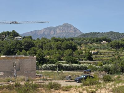 Vue d'un chantier avec une structure en blocs de béton et une grue, encadré par une végétation méditerranéenne luxuriante, des collines en terrasses et la montagne du Montgó sous un ciel clair.