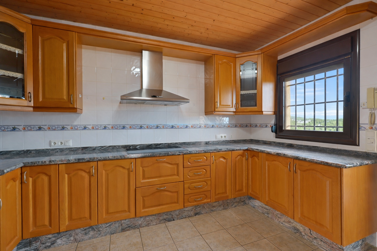 Cuisine aménagée avec des meubles en bois, des plans de travail en granit et un plafond en lattes de bois. Fenêtre grillagée avec vue extérieure. Cuisine aménagée avec des meubles en bois, des plans de travail en granit et un plafond en lattes de bois. Fenêtre grillagée avec vue extérieure.
