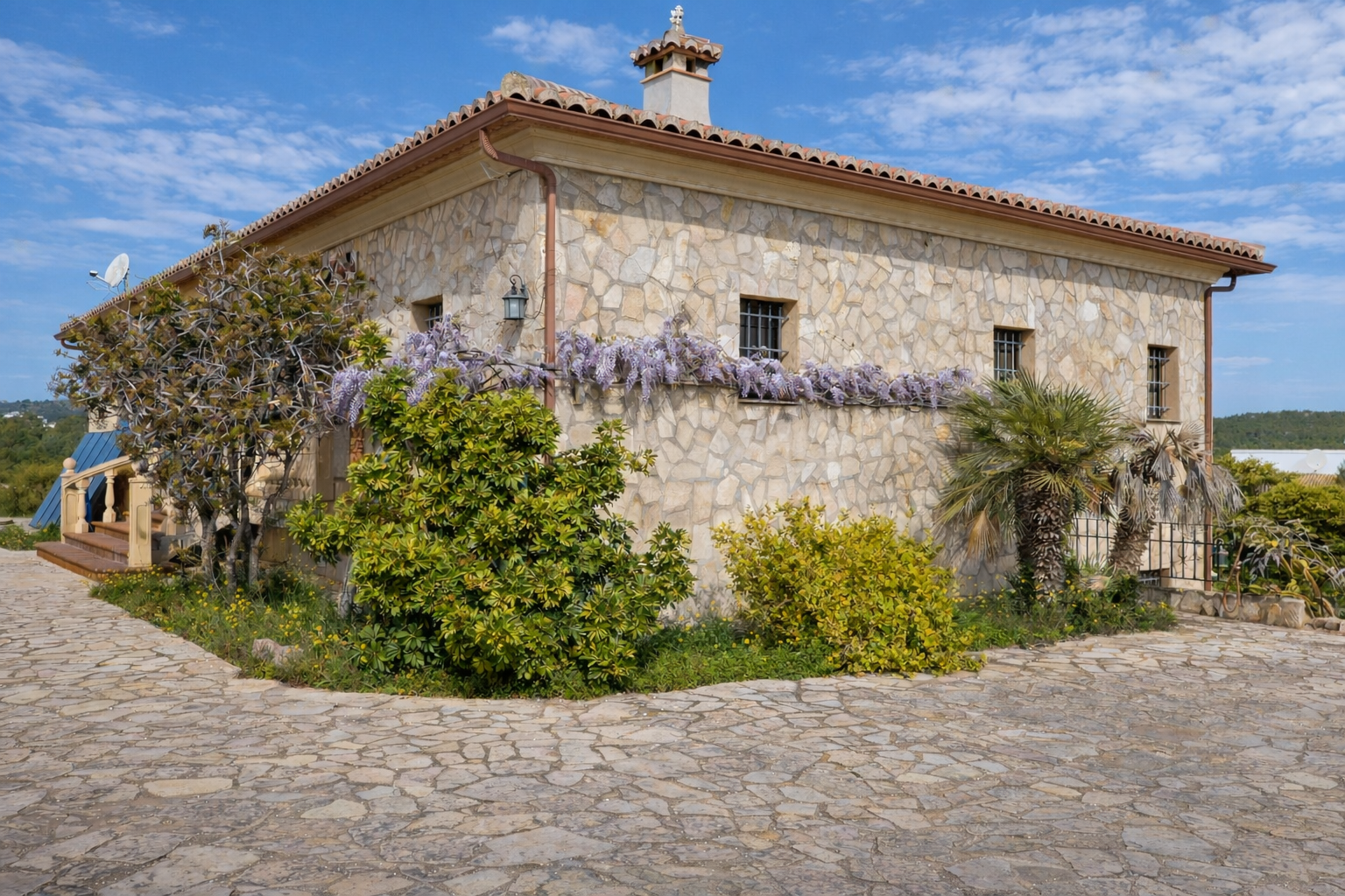 Façade en pierre rustique avec toit en tuiles, ornée de glycines violettes sous les fenêtres grillagées. L'allée est pavée de pierres irrégulières. Façade en pierre rustique avec toit en tuiles, ornée de glycines violettes sous les fenêtres grillagées. L'allée est pavée de pierres irrégulières.