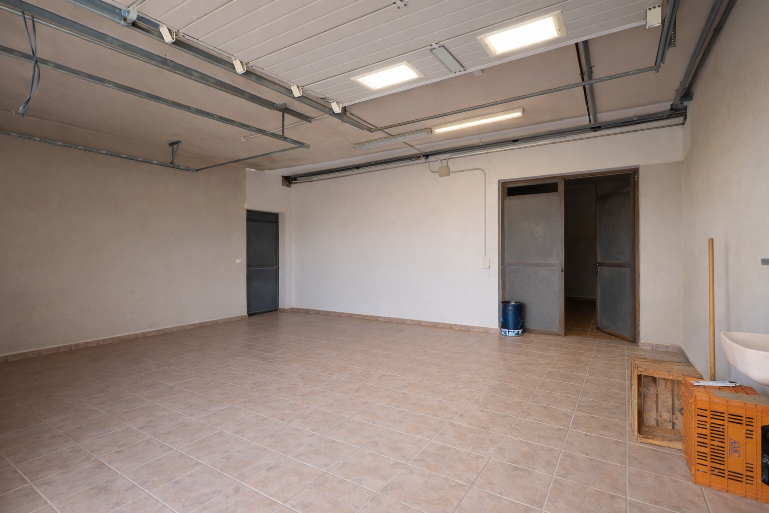 Grand garage ou espace utilitaire avec carrelage clair, éclairage de plafond industriel et multiples points d'accès. Grand garage ou espace utilitaire avec carrelage clair, éclairage de plafond industriel et multiples points d'accès.