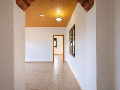 Couloir intérieur avec carrelage clair et murs blancs texturés. Plafond en bois clair avec luminaire central et détails sculptés à l'entrée.