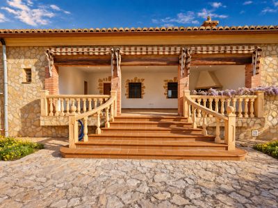 Entrée principale avec vaste naya au sol en terre cuite, façade en pierre naturelle et escalier à balustrade classique. Une cheminée, de la glycine et un panneau solaire sont visibles.