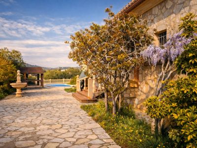Façade en pierre de la finca avec glycine en fleurs. Un chemin dallé mène à la piscine et à une pergola en bois, avec une fontaine en pierre dans le jardin.