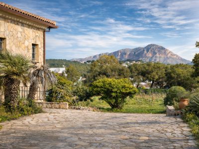 Façade en pierre rustique de la finca avec palmiers, allée pavée et vues sur le Montgó, entourée de vignobles et végétation méditerranéenne.