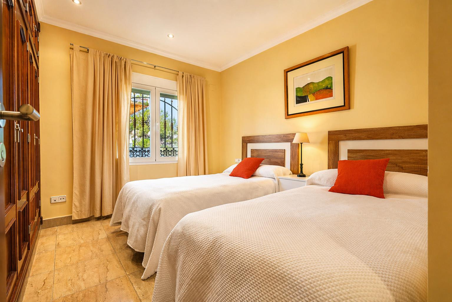 Chambre double avec lits jumeaux, têtes de lit en bois et coussins orange. Elle dispose d'une armoire intégrée, d'un sol carrelé et d'une fenêtre à barreaux.